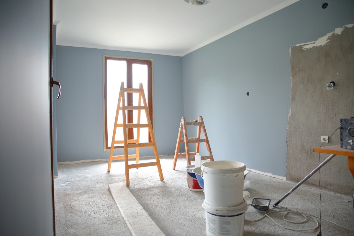 A photo of a room that is being redecorated. The walls are grey whereas the floor is carpeted and it is white/very light grey in colour. There are two sets of ladders and paint buckets.